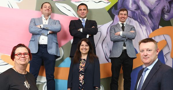 A photograph of six people, four men and two women, stood in smart work clothing in front of a large, colourful orange, purple and green mural. The group are stood in a horseshoe shape but all are facing the camera and smiling